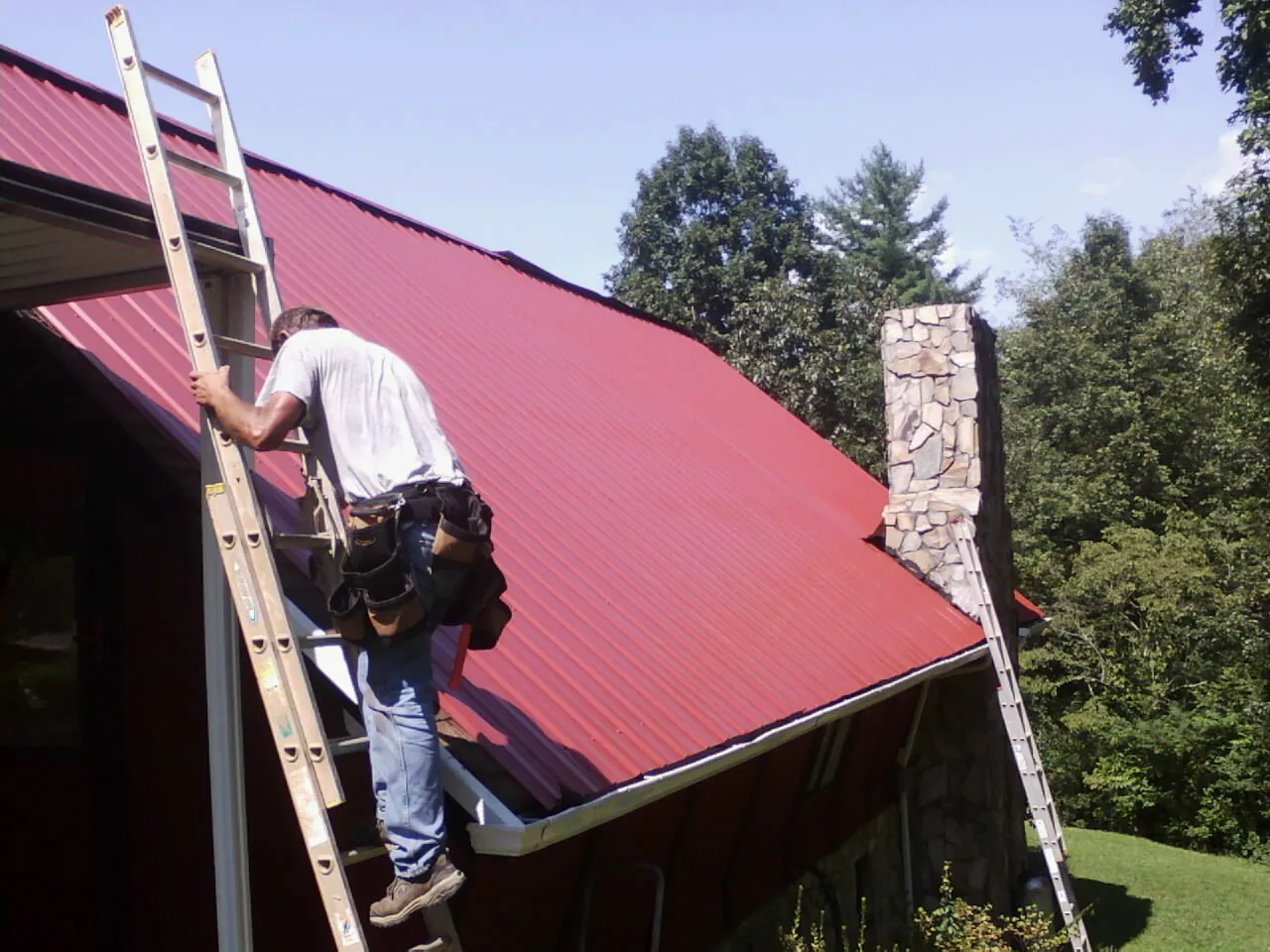 Roofer on a ladder beside a red metal roof and stone chimney — Soffit & Fascia Repair by Roofing Brothers in Armuchee
